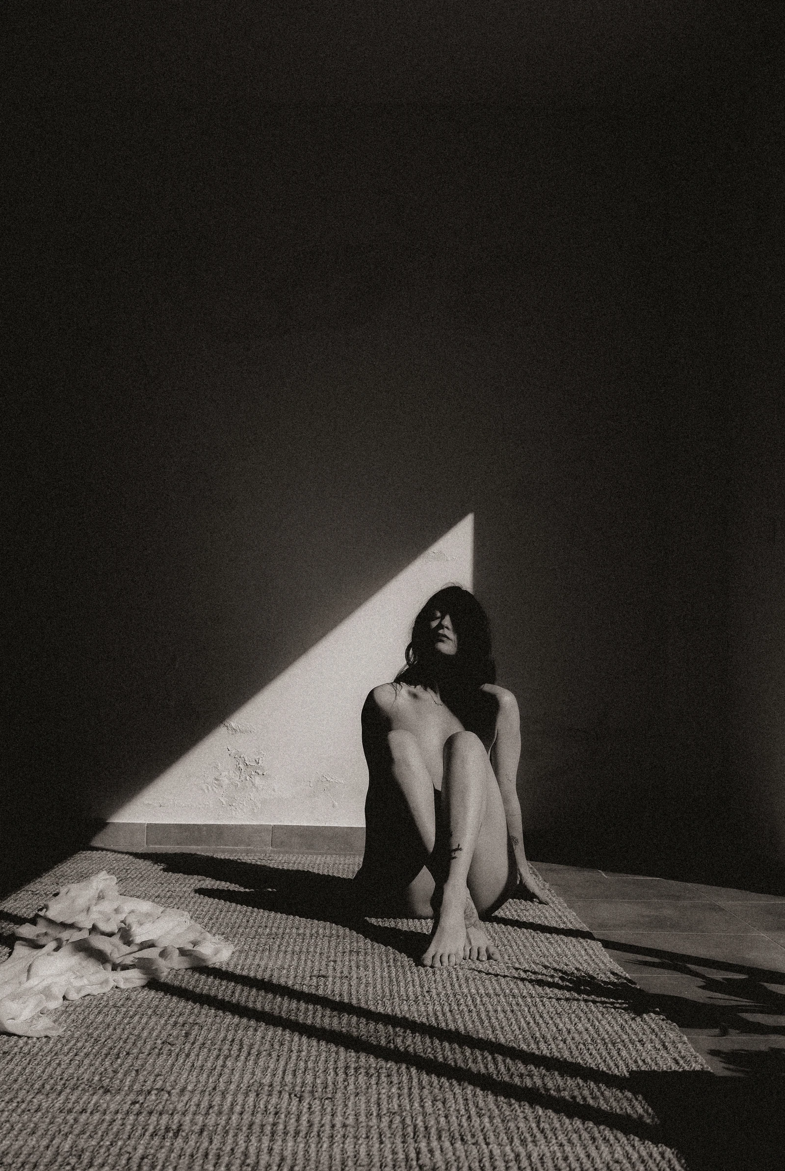 A seated figure draws their knees close in a triangle of sunlight on a wall.