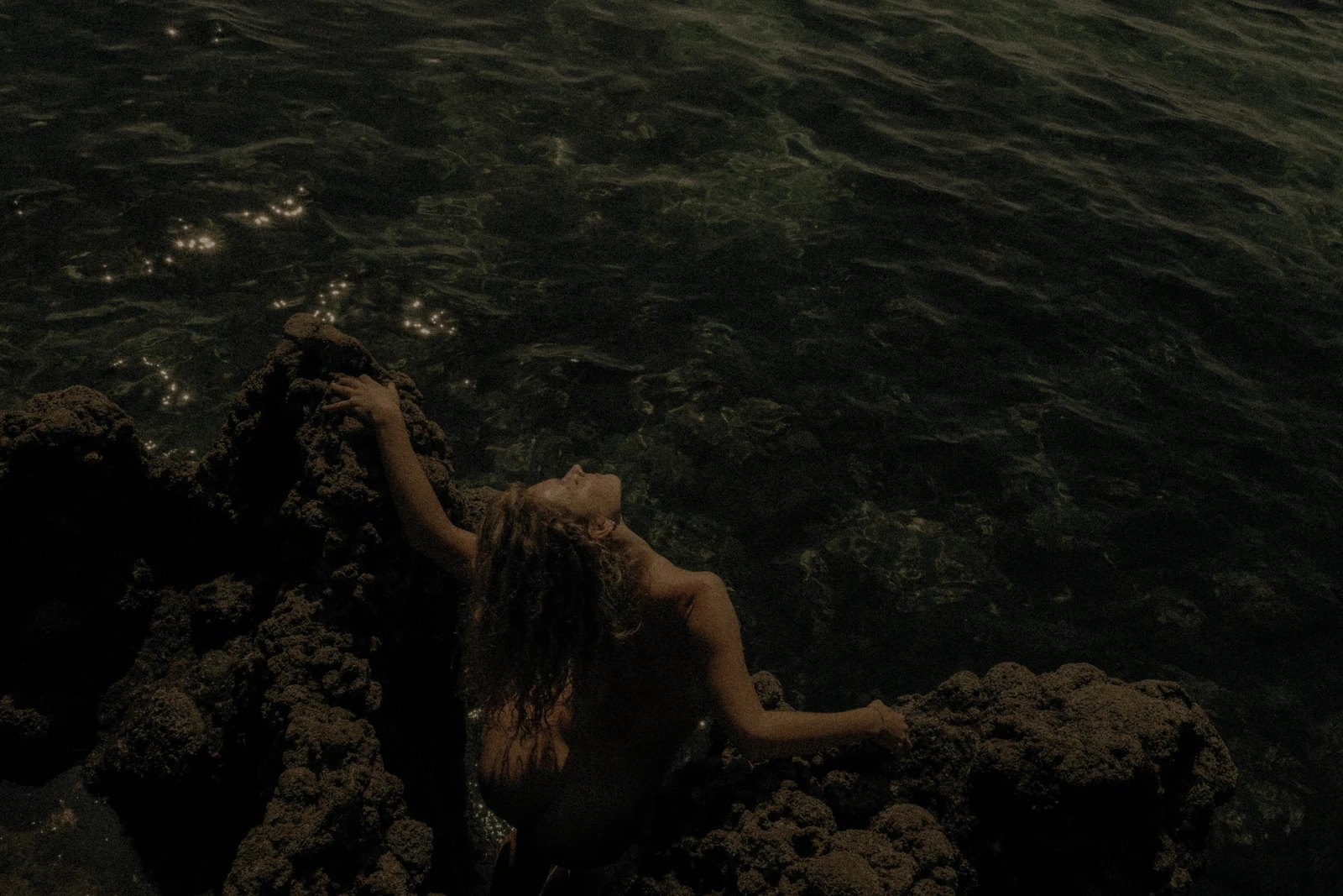 A figure holds the rocks at the water’s edge, face lifted towards the light.